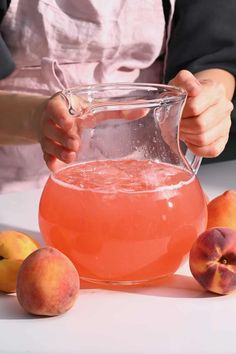 a person holding a pitcher filled with liquid next to peaches on a white table