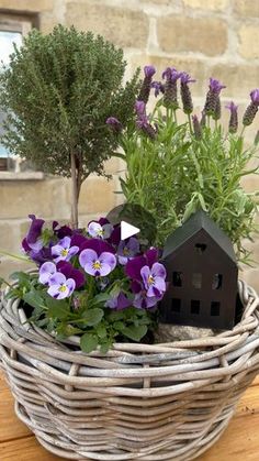 purple flowers in a wicker basket next to a house shaped planter on a wooden table