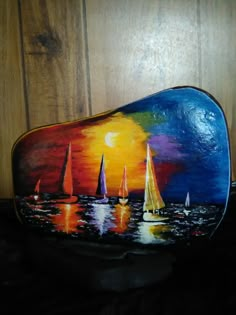 a painted rock with sailboats on the water at sunset in front of a wooden wall