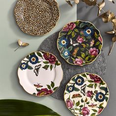 three plates with flowers and butterflies on them sitting next to a green leafy plant