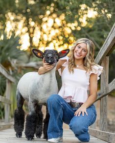 a woman kneeling down next to a sheep on a wooden bridge with trees in the background