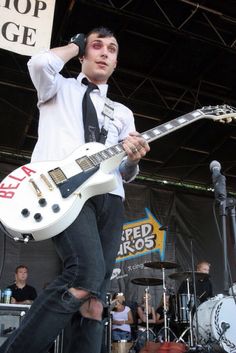 a man in white shirt and black tie playing guitar on stage with other people behind him