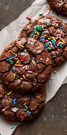 three chocolate cookies with sprinkles and colored candies on top of parchment paper