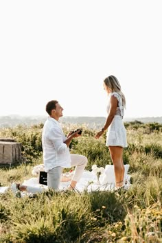 a man kneeling down next to a woman on top of a grass covered field while holding a cell phone