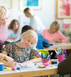 two women sitting at a table working on some art projects with other people in the background