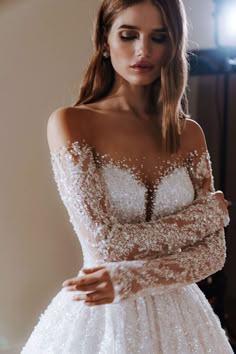 a woman wearing a white dress with long sleeves and beading on the shoulders is posing for