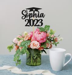 there is a vase with flowers and a graduation cake topper next to a cup