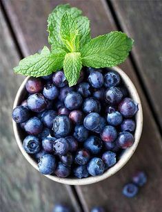 a small bowl filled with blueberries on top of a wooden table next to mint leaves