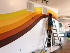 a woman standing on a ladder painting a wall with rainbow stripes and white shutters