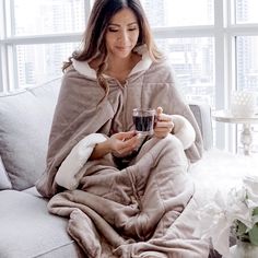 a woman sitting on a couch wrapped in a blanket and holding a cup with both hands