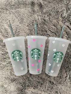 three starbucks cups with straws in them sitting on a carpeted surface, one has pink hearts and the other is green