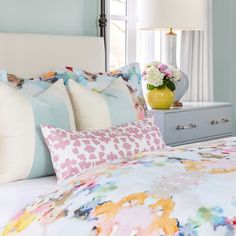 a bed with colorful sheets and pillows in a bedroom next to a dresser filled with flowers