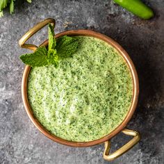 a wooden bowl filled with green sauce next to cucumbers and mint leaves on a gray surface