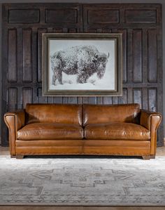 a brown leather couch sitting in front of a wooden paneled wall with a buffalo painting above it