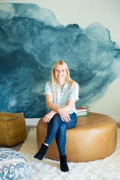 a woman sitting on top of a leather ottoman in front of a painting and rug