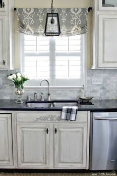 a kitchen with white cabinets and black counter tops in front of a window that has roman shades