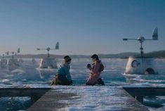 two people are sitting on a pier looking at something in the distance while another person is standing near them