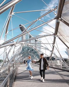 two people walking down a walkway under a blue sky with buildings in the background,