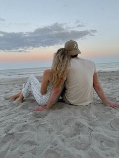 a man and woman sitting on top of a sandy beach next to the ocean at sunset