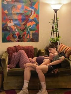 two people sitting on a couch in front of a painting and lamp, one holding the other's head