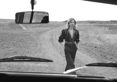 a woman walking down a dirt road in front of a car with a rear view mirror