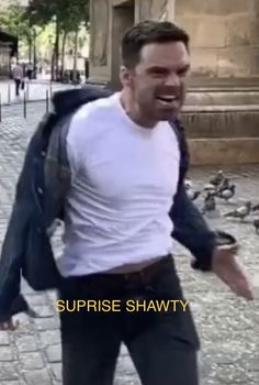 a man in white shirt and black jacket walking on street with pigeons behind him at daytime