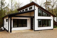 a white house with black trim on the roof and windows, sitting in gravel area next to trees