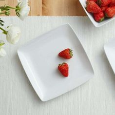 strawberries are placed in square white dishes on a place mat next to some flowers