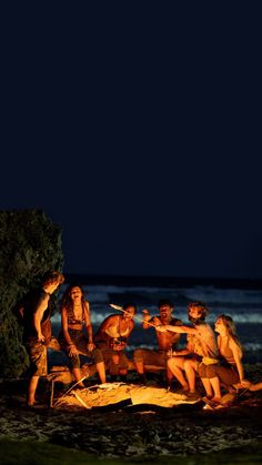 a group of people sitting around a campfire on the beach in front of the ocean