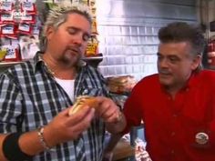 two men standing in front of a counter and one is holding a hot dog with ketchup on it