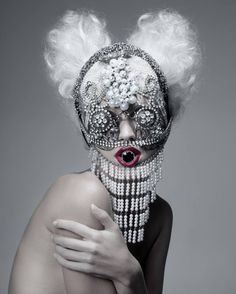 a woman with white hair wearing a mask and pearls on it's face, posing in front of a gray background