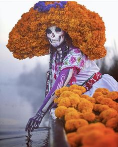 a woman with flowers on her head sitting in front of some water and orange flowers