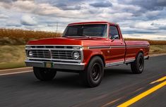 an old red pickup truck driving down the road with cloudy skies in the back ground