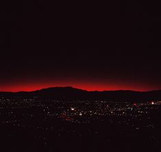 the sky is dark and red as seen from an overcast city at night with mountains in the distance