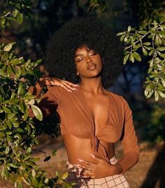 a woman with an afro standing in front of some trees and bushes wearing a brown top