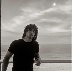 a man with dreadlocks standing on a balcony near the ocean looking at the camera