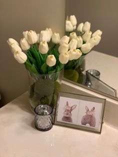 a vase filled with white tulips next to a framed photo on a bathroom counter