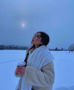 a woman is standing in the snow holding a cup and looking up at the sky