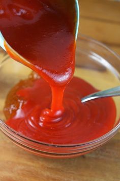 a spoon pouring ketchup into a glass bowl on top of a wooden table