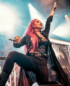 a woman with pink hair and black clothes on stage holding her arms in the air