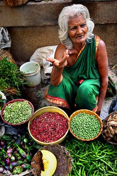 an older woman sitting in front of bowls of vegetables and fruits on the ground with her finger up