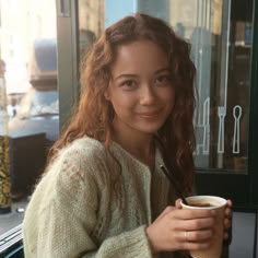 a woman sitting at a table with a cup of coffee in her hand and looking into the camera