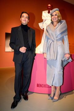 a man and woman standing next to each other in front of a pink wall with a sculpture behind them