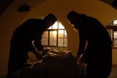 two men standing over a bed in a dimly lit room, one is holding the head of another man