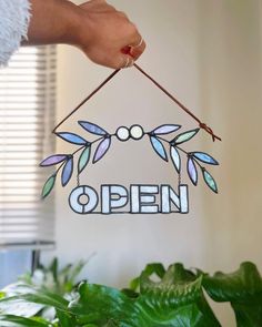 a hand holding an open sign in front of a potted plant with green leaves