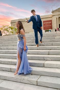 a woman in a long blue dress standing on steps with a man walking behind her