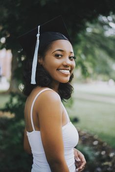 a woman wearing a graduation cap and gown smiling at the camera with trees in the background