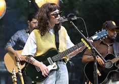 a woman singing into a microphone while playing an electric guitar and another man plays the guitar behind her