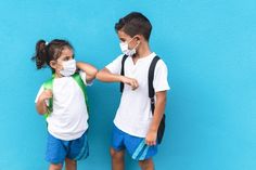 two children wearing face masks and backpacks standing against a blue wall with their arms around each other