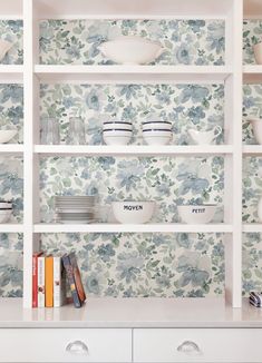 a white book shelf filled with plates and bowls on top of each other in front of a floral wallpaper